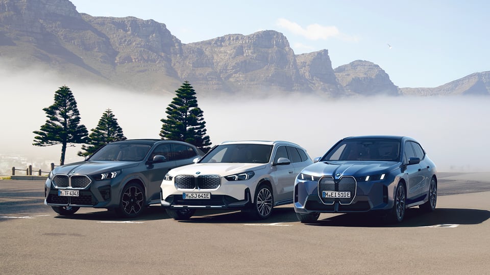 Three BMW vehicles parked on an open road with misty mountains and pine trees in the background