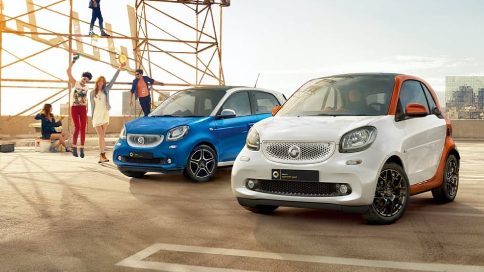 Two small smart cars, one white with orange accents and one blue, parked on rooftop with five people having fun nearby
